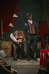 High African man standing with little servant boy carrying heavy suitcases on his back over dark retro circus backstage background. Concept of circus, theater, performance, show, retro and vintage