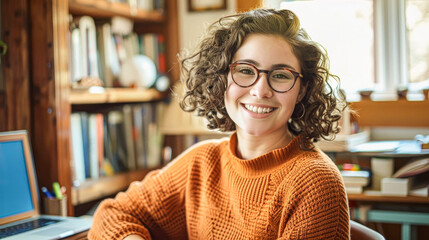 A woman with curly hair and glasses is smiling at the camera. She is sitting at a desk with a laptop and a stack of books. Concept of comfort and relaxation
