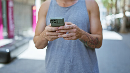 Young tattooed man using smartphone on a sunlit city street, creating a modern urban vibe.