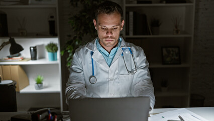 Hispanic male doctor in lab coat working on laptop at nighttime in clinic office, evoking...