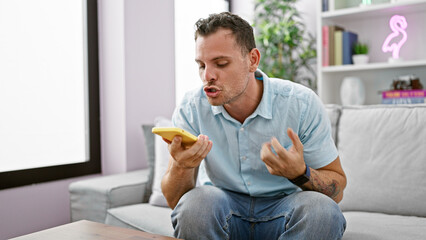 Fototapeta na wymiar Angry young hispanic man with a beard in casual attire using a smartphone in a modern living room.