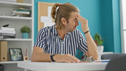 A stressed man with long blond hair in a striped shirt expresses worry in a modern office setting.