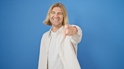 Handsome blond man pointing and smiling against a solid blue background, personifying confidence...