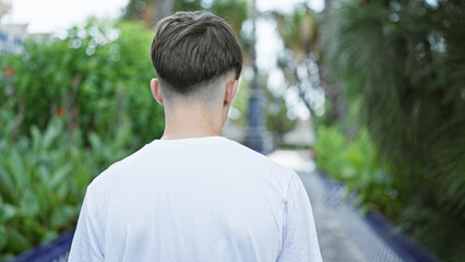 Rear view of a young caucasian man in a white shirt standing outdoors amidst lush greenery on a sunny day.
