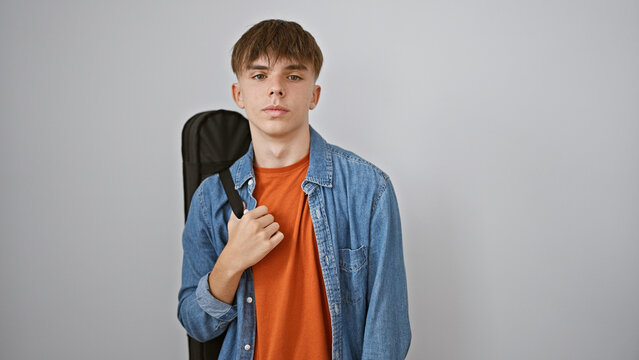 A young caucasian male teenager with blonde hair, carrying a guitar case, poses against a white wall.