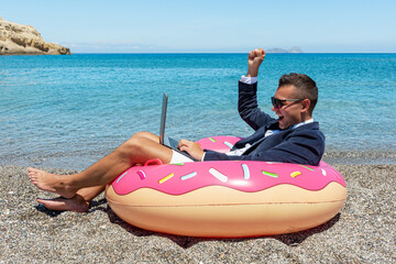 Happy businessman with laptop on inflatable donut on tropical beach.