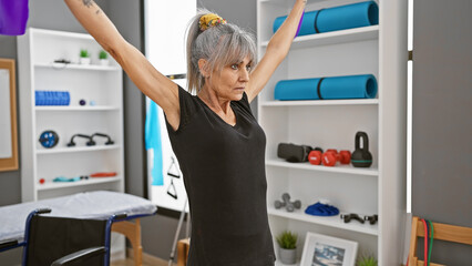 A mature hispanic woman exercising with outstretched arms in a rehab clinic's gym, indoors.