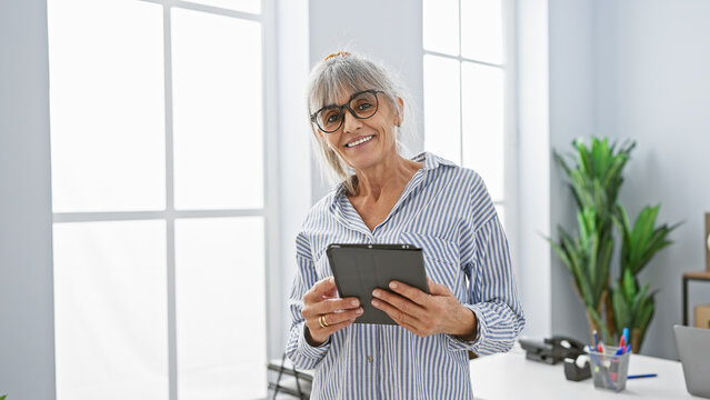 A smiling mature woman with grey hair and glasses standing in a modern office holding a tablet.