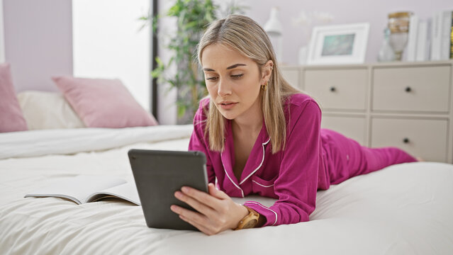 A young woman in pink pajamas using a tablet while lying on a bed in a well-lit bedroom.