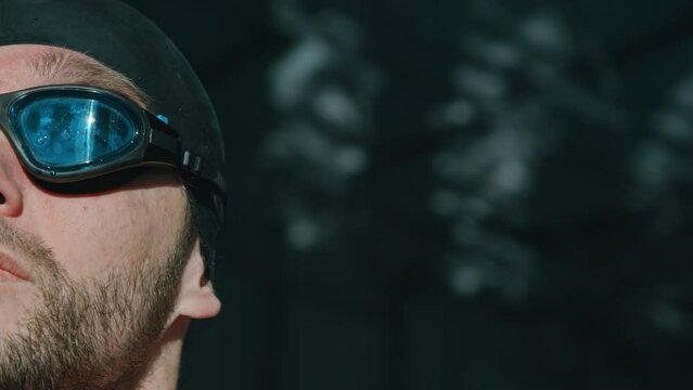 Extreme close-up half face portrait footage of confident, ambitious young Caucasian swimmer with stubble, beard, in black rubber cap and blue goggles mentally preparing for competition in pool