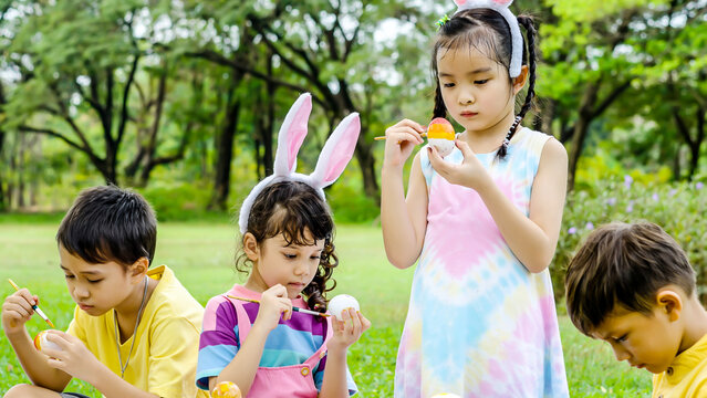 Happy group children in park, cute Asian girl and boy with mix race friend wear bunny ears, paint egg with paintbrush together on green grass in garden. Kids celebrate Easter holiday outdoor