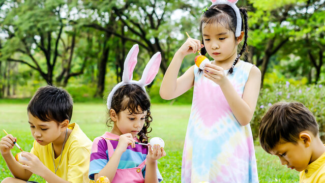 Happy group children in park, cute Asian girl and boy with mix race friend wear bunny ears, paint egg with paintbrush together on green grass in garden. Kids celebrate Easter holiday outdoor