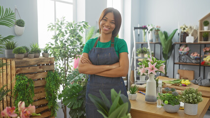 Smiling woman with crossed arms wearing an apron in a lush flower shop interior.