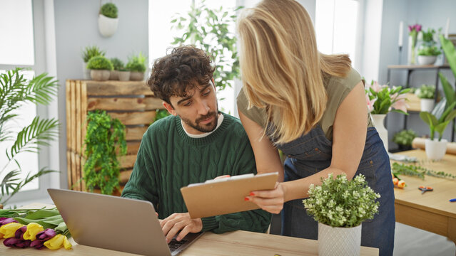 Man and woman collaborate in a flower shop with laptops, plants, and bouquets against an indoor green background.