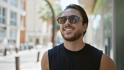Young hispanic man wearing sunglasses looking to the sky smiling at street