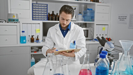 Young hispanic man scientist writing on notebook sitting at laboratory