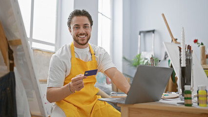 Young hispanic man artist shopping with credit card and laptop smiling at art studio
