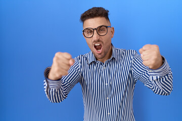 Hispanic man with beard wearing glasses angry and mad raising fists frustrated and furious while shouting with anger. rage and aggressive concept.