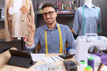 Hispanic man with beard dressmaker designer working at atelier showing and pointing up with fingers number three while smiling confident and happy.