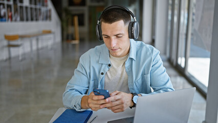 A focused young man listens to headphones while using a smartphone and laptop in a library setting.