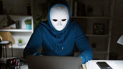 A incognito man in a hoodie works intently on a laptop at night in a home office. - obrazy, fototapety, plakaty