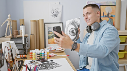 Smiling young man with headphones using smartphone in art studio surrounded by paintings and supplies.