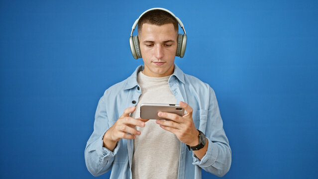 Hispanic man in casual attire using smartphone with headphones against blue background outdoor