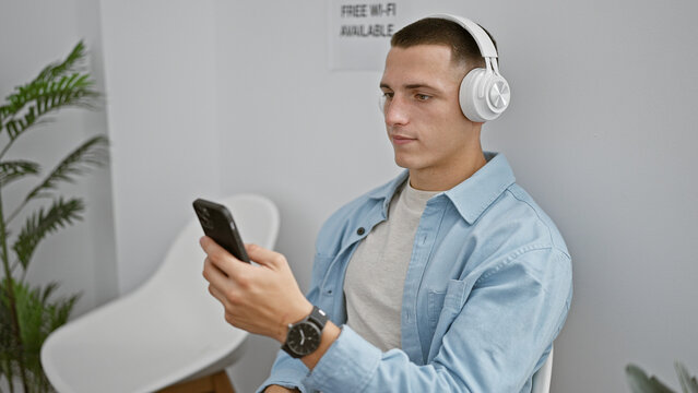 A young hispanic man wearing headphones uses a smartphone in an indoor room with a white chair and plant.
