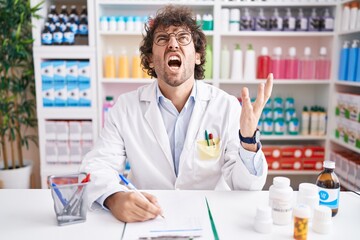 Hispanic young man working at pharmacy drugstore crazy and mad shouting and yelling with aggressive expression and arms raised. frustration concept.