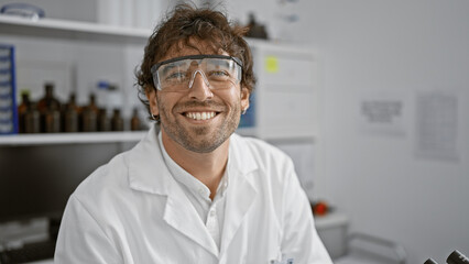 A cheerful hispanic man with a beard and green eyes, wearing safety goggles and a lab coat, stands...
