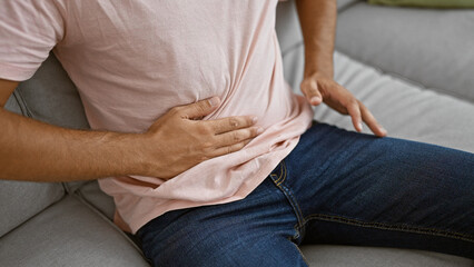A young hispanic man experiencing abdominal pain sits on a sofa at home, capturing a moment of...