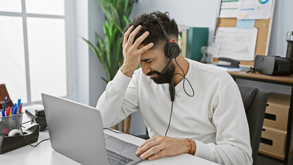 A stressed hispanic man with a beard wearing headphones looks troubled at work in a modern office...