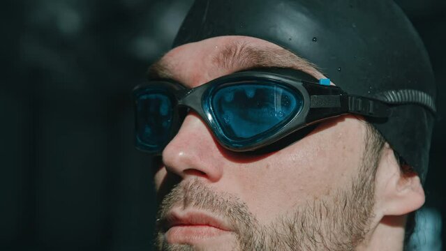 Extreme close-up portrait shot of confident, ambitious young Caucasian sportsman with stubble and beard, in black rubber cap putting on goggles in pool, looking away then at camera