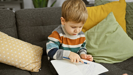 A focused blonde toddler boy is doodling in a notebook while sitting on a couch inside a cozy...