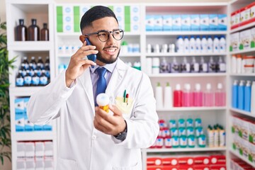 Young latin man pharmacist holding pills bottle talking on smartphone at pharmacy
