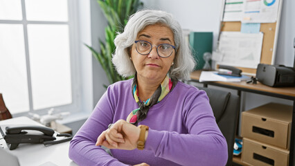 Mature woman in glasses checking time in modern office, evoking time management concepts.