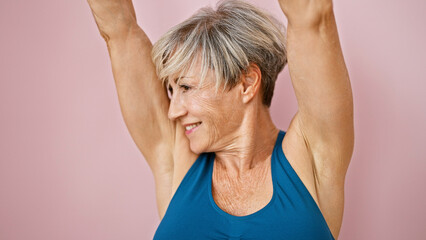 A smiling mature woman with short grey hair in a blue tank top stretching her arms against a pink...