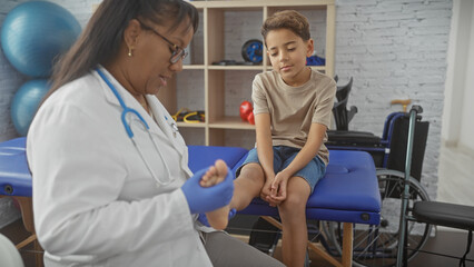 Obraz premium A woman therapist examines a boy's foot in a physiotherapy clinic's rehab room, suggesting a pediatric healthcare setting.