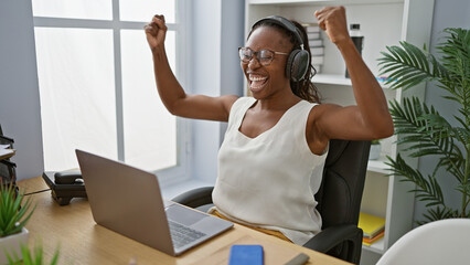 An adult african american woman celebrates with raised arms at her workplace, wearing headphones...