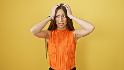 A perplexed young woman in an orange top clutching her head against a solid yellow background,...