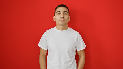 A young, handsome hispanic man in a white shirt stands against a vibrant red wall.
