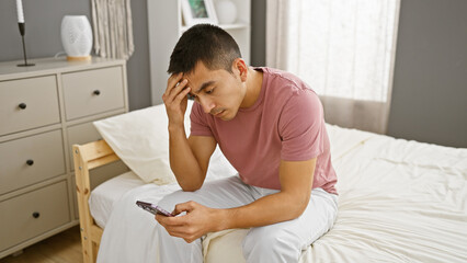 Pensive young hispanic man with phone sitting on bedroom bed in a modern apartment interior.