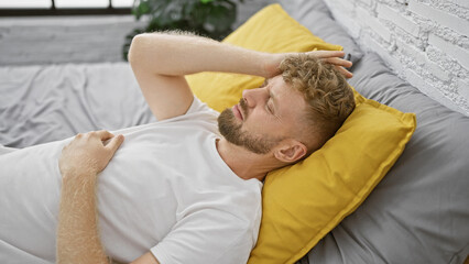 A relaxed young man with a beard and blue eyes resting in a bedroom with yellow pillows and white brick wall background.