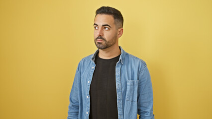Handsome hispanic man with beard in casual denim jacket against yellow background, looking aside...