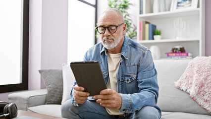 Mature man in denim using tablet on couch in bright living room
