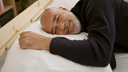 Smiling middle-aged bald man with grey beard relaxing in bedroom, giving a sense of calm and...