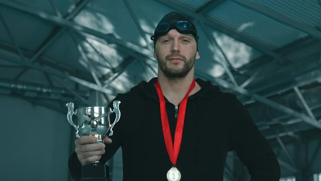 Medium close-up shot of young Caucasian male swimmer in black cap, goggles, zip hoodie, with gold medal on neck and trophy cup in hand, posing for photos in pool and smiling for camera