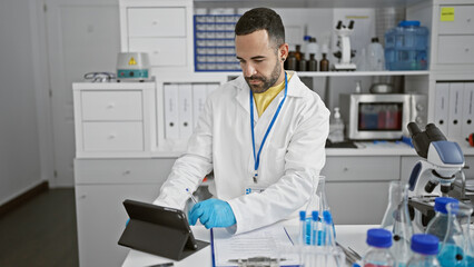 Fototapeta na wymiar Hispanic man in lab coat using tablet in laboratory setting with equipment and microscope