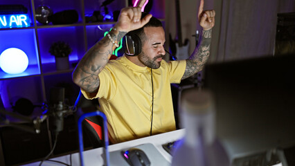 A cheerful hispanic man with tattoos celebrates while gaming at night in a well-lit room with modern equipment.