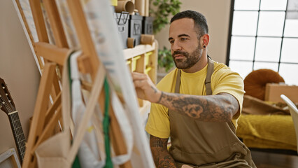 Handsome hispanic man with tattoos painting on canvas in a bright art studio